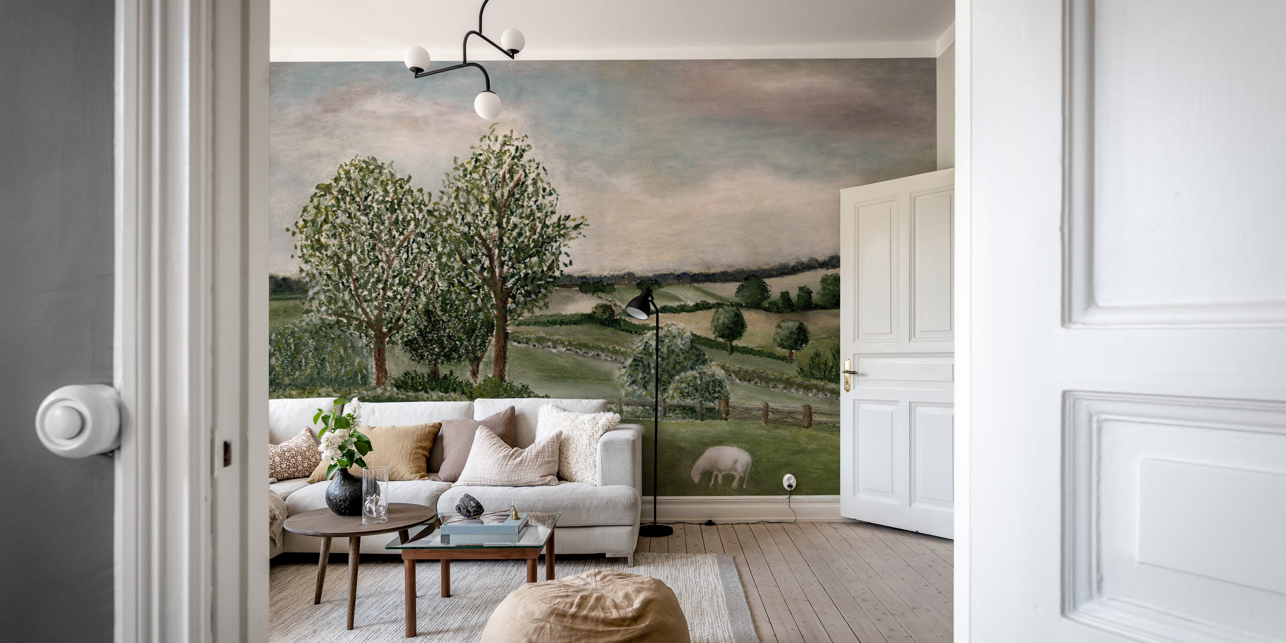 pastoral-landscape-mural-wallpaper-living-room Pastoral landscape mural wallpaper with grazing sheep and rolling green fields in a living room with a cream sofa and jute rug